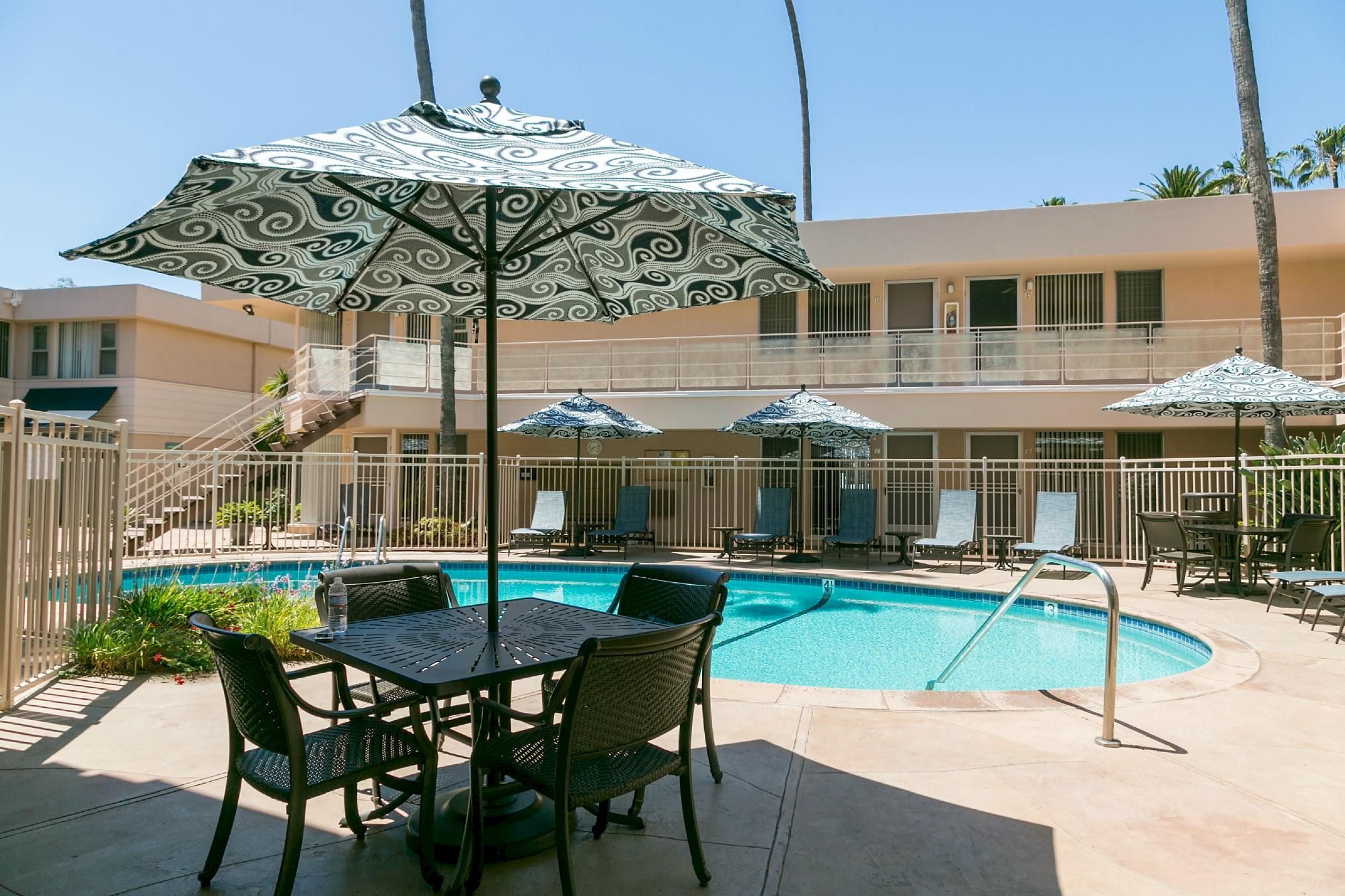 A sunny poolside patio with a rectangular pool, outdoor tables and umbrellas, and a two-story motel-style building in the background.