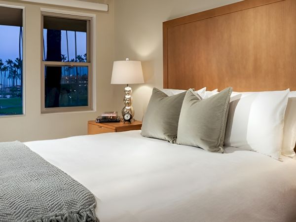 A modern hotel bedroom with a large bed, white linens, gray pillows, wooden headboard, matching nightstands, and lamps; calm, tidy vibe.