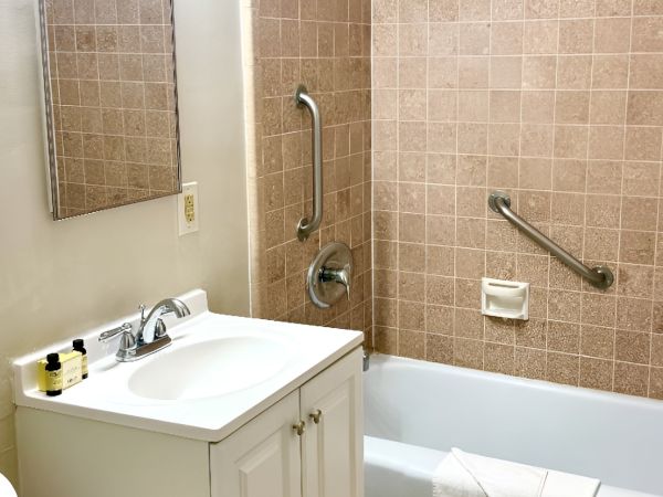 A small bathroom with a white vanity, sink, mirror, and a tub/shower combo with beige tile and safety grab bars. End.