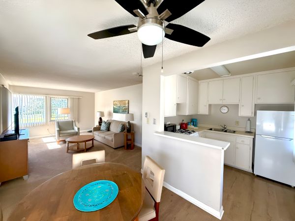 A cozy open-plan living room and kitchen with white cabinets, a ceiling fan, and a small dining table, blending seating area and breakfast bar.