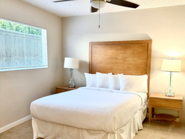 A tidy bedroom with a queen bed, white bedding, wooden headboard, two matching nightstands, lamps, a ceiling fan, and a window with blinds.