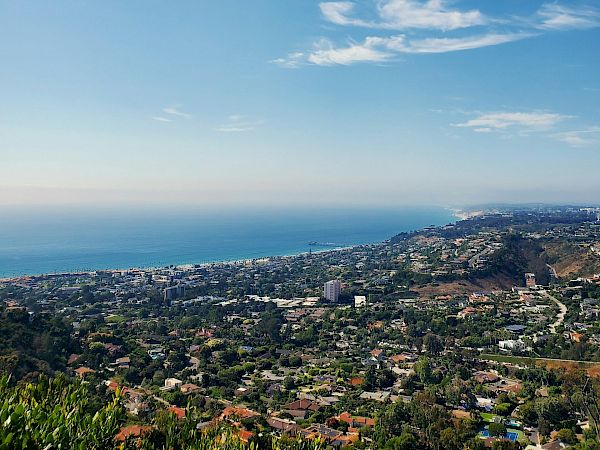 A scenic coastal valley with a sprawling town, hills, and a calm blue sea on the horizon under a clear sky.