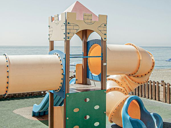 A beige playground castle with orange slides and tubes, set on green turf by the sea, under a clear blue sky.