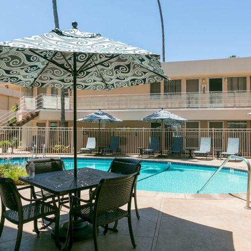 A sunny poolside courtyard with a rectangular pool, lounge chairs, and patterned umbrellas around a small seating table, two-story beige hotel in background.