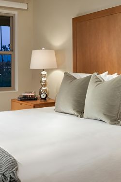 A cozy hotel bedroom with a large bed, white linens, gray throw, three pillows, wooden headboard, and matching nightstands with lamps.