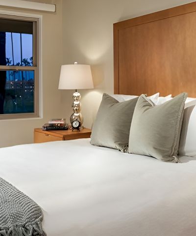 A cozy hotel bedroom with a large bed, white linens, gray throw, three pillows, wooden headboard, and matching nightstands with lamps.