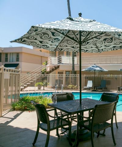 A sunny hotel pool area with lounge chairs, umbrellas, and a table set on a patio surrounding a sparkling blue pool.