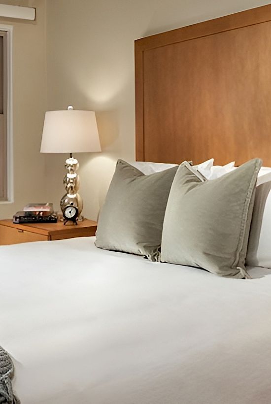 A neatly made hotel bedroom with a large bed, white linens, gray throw, wooden headboard, two bedside tables, and matching lamps.