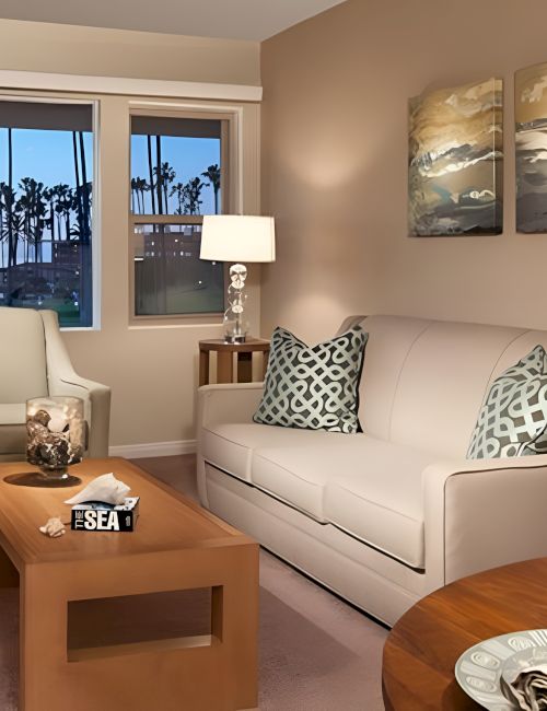 Modern hotel-style living room with a beige sofa, matching armchairs, wooden coffee table, and round dining table. Large windows overlook palm trees. Cozy lamps, throw pillows, and tasteful artwork complete the space.