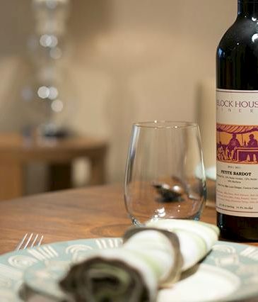 A cozy table setting with a bottle of red wine, two glasses, a rolled napkin on a plate, and a seashell decor item in a stylish room.