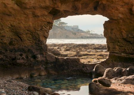 A rocky coastal arch opens to a calm sea with a small pool at the foreground, framed by rugged cliffs.