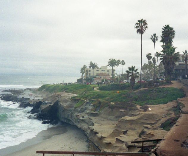 A rocky coastal scene with waves crashing along a shore, palm trees in the distance, and a cloudy sky at a calm, breezy moment.